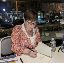 ANN WALLACE SEATED, SIGNING A BOOK